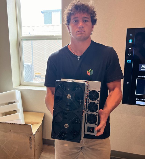 The image shows a man standing next to a window, holding two computer fans or cooling units. He appears to be posing for a photo while displaying his equipment. The background of the image features a TV screen mounted on the wall above him. There is also an open cardboard box in front of the man and another one behind him.