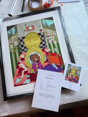 A framed colorful illustration of a yellow character alongside a card and an information sheet rests on a surface with packing tape visible.