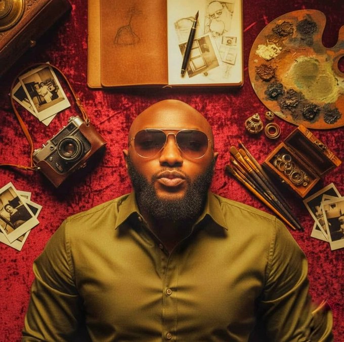 An overhead portrait of a bald man with a beard wearing sunglasses and an olive shirt, surrounded by artistic items including a camera, sketchbook, paint palette, and photos on a red surface.