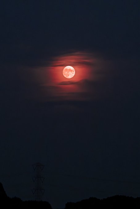 A glowing red full moon, likely a blood moon, shines through wispy clouds in a dark night sky above silhouetted hills and a power line tower.