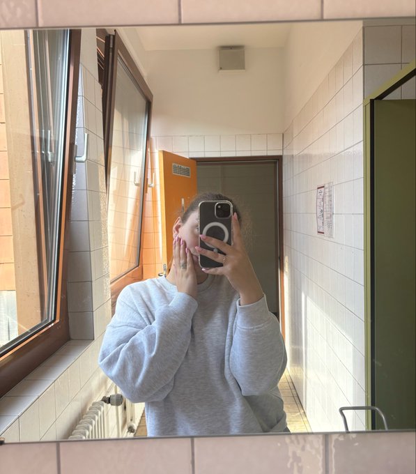 A mirror selfie taken in a tiled bathroom, showing a person wearing a gray sweatshirt holding a smartphone to take the photo while touching their face.