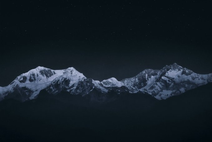 Snow-capped mountain peaks, likely representing Kangchenjunga—the world's third-highest mountain—rise against a dark, starry night sky, creating a serene and majestic atmosphere.