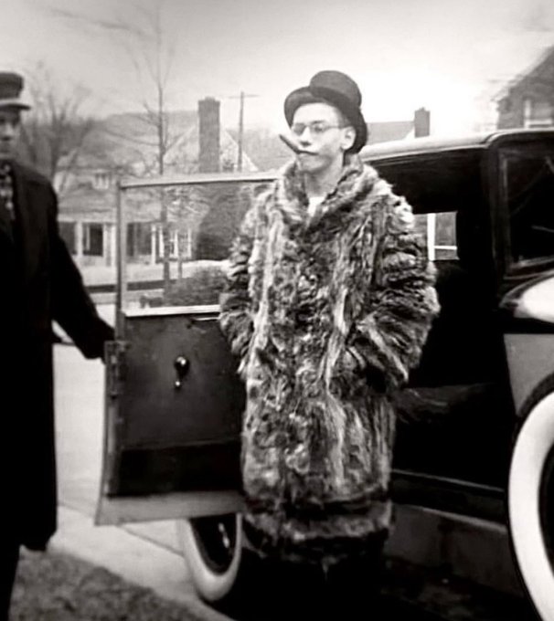 A vintage black and white photo of a man wearing a top hat, round glasses, and a large fur coat, standing next to an open car door with a cigar in his mouth.