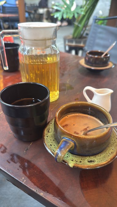A café table holds a black mug, a cup of coffee with a spoon, a small cream pitcher, and a tall glass pitcher of golden liquid.