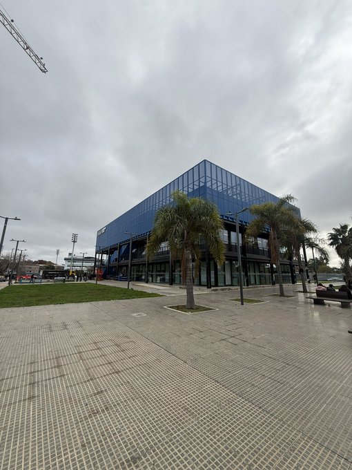 The image shows an empty plaza with a large blue building featuring a glass facade that reflects the cloudy sky above. The plaza has a sidewalk running through its center, where several palm trees can be seen. There are also benches placed around the area for people to sit and enjoy the surroundings. A construction crane is visible in the background, indicating ongoing development or renovation work nearby.