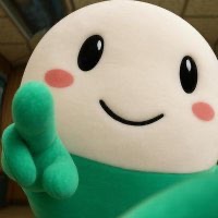 A white cartoon mascot character with pink cheeks and a green outfit giving a peace sign.