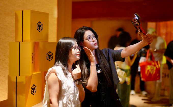 Two young women taking a selfie at what appears to be a fan event or convention, standing in front of a stack of yellow boxes featuring a cube logo.