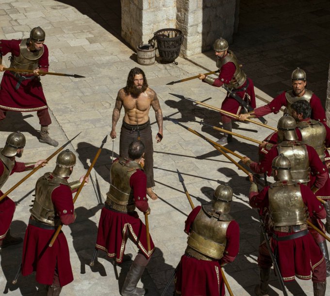 A shirtless, bearded man stands defiantly surrounded by armored soldiers pointing spears at him in a sunlit stone courtyard. The image metaphorically illustrates the post's argument that Polymarket has undermined its own strong first-half momentum by pivoting to compete with traditional markets, essentially self-sabotaging by stepping into a battlefield (stocks, crypto, commodities) where it faces stronger, entrenched competitors.