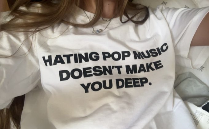 A person wears a white t-shirt with bold black text reading "HATING POP MUSIC DOESN'T MAKE YOU DEEP." along with a silver pendant necklace.