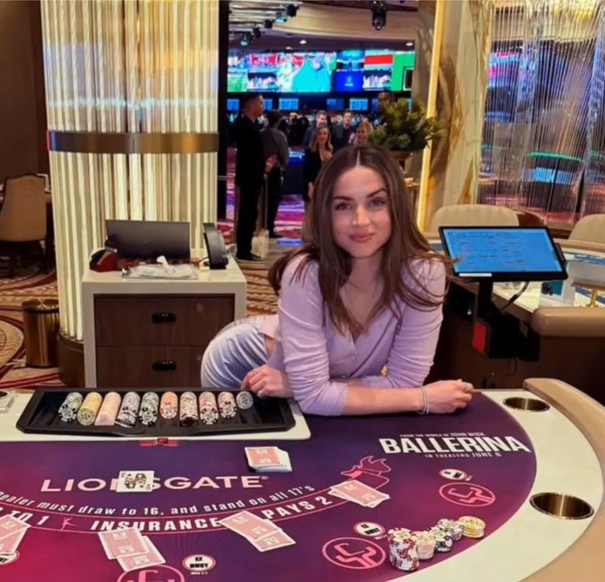 Young woman posing at a casino table with sushi rolls in front of her; the table reads 'BALLERINA' and 'LION GATE PAYS 2' with the instruction 'Dealer must draw to 16, and stand on all 17's'.