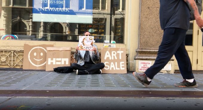Street photography shot of a person sitting cross-legged on a sidewalk against cardboard signs reading 'H2M' with a smiley face logo and 'ART SALE,' holding up artwork, while a passerby's legs cross the frame.