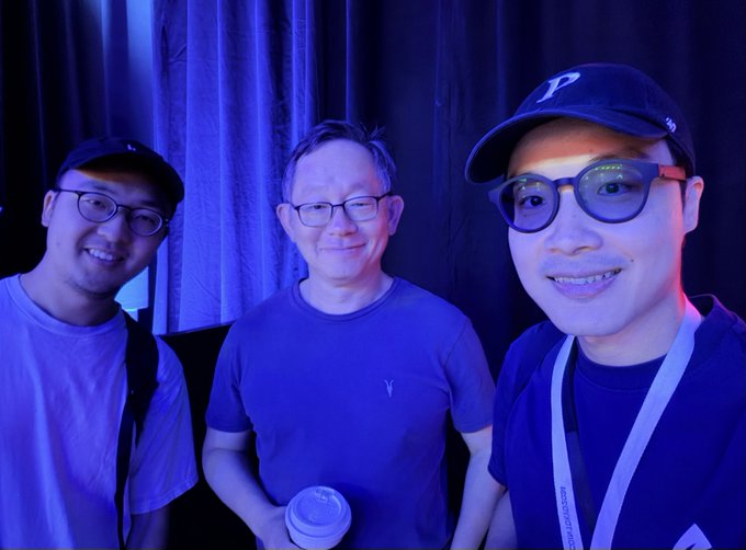 Three men smile for a selfie in a dimly lit room with blue lighting; the man in the center wears glasses and a blue shirt while holding a white cup, and the man on the right wears glasses, a blue shirt, and a dark baseball cap with a white 'P' logo.