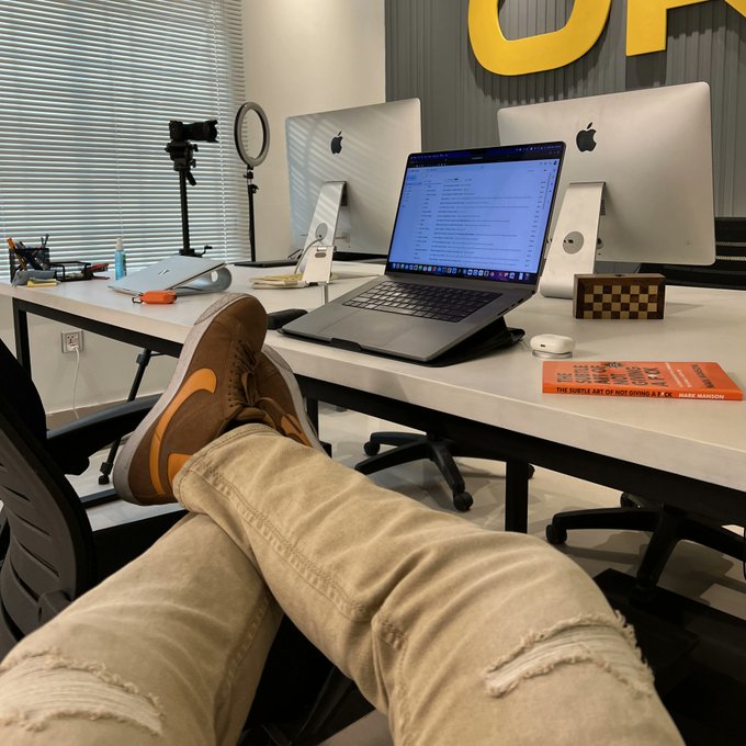 A first-person view shows someone with their feet propped up on a desk featuring a MacBook, two Apple monitors, a ring light, and a book titled 'The Subtle Art of Not Giving a F*ck' by Mark Manson.
