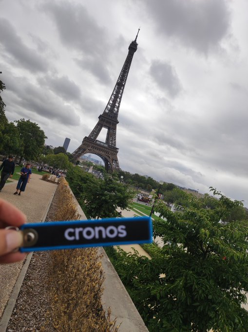 A photo taken at the Eiffel Tower in Paris showing a hand holding a keychain with the text 'cronos' in the foreground, with the iconic tower and cloudy sky in the background.