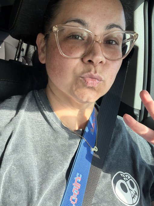 A woman with glasses and dark hair takes a selfie in a car, wearing a gray t-shirt with a skull design, a gold necklace, and a seatbelt with a 'Chain' branded strap.