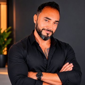 A portrait of a bearded man with a chest tattoo, wearing a black button-down shirt with his arms crossed, wearing a wristwatch against a blurred background.