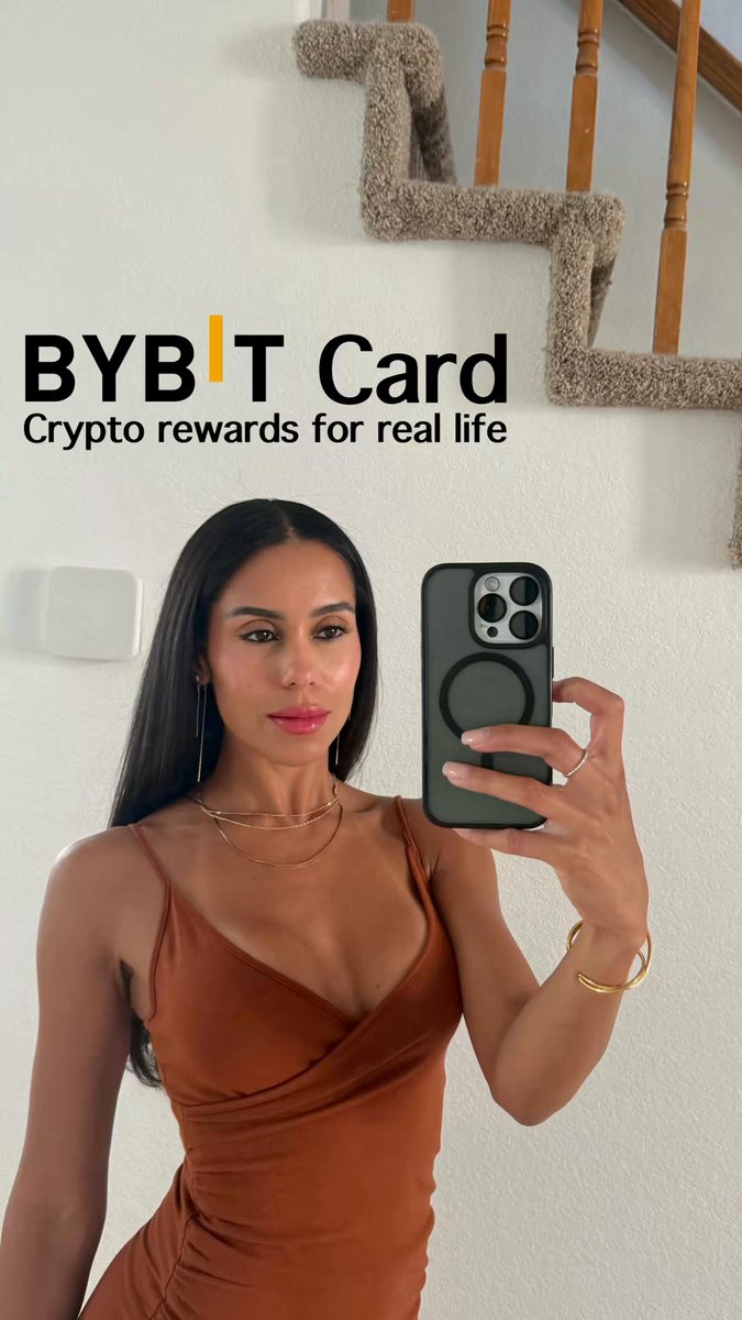 The image features a woman taking a selfie with her cell phone, holding it up against a white wall. She appears to be wearing an orange dress or top. The background of the photo includes a staircase, which adds depth to the scene. There is also text in the image that reads "Bybit Card", suggesting that the photo might be related to a Bybit card or credit card.