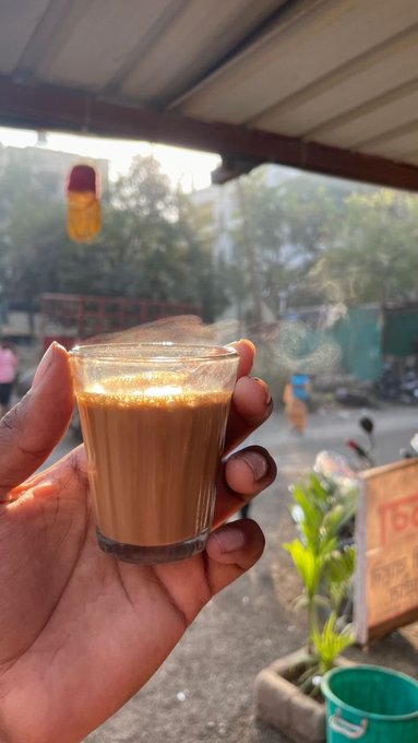 A hand holding up a small glass of chai tea outdoors, with a sunny street scene visible in the background.