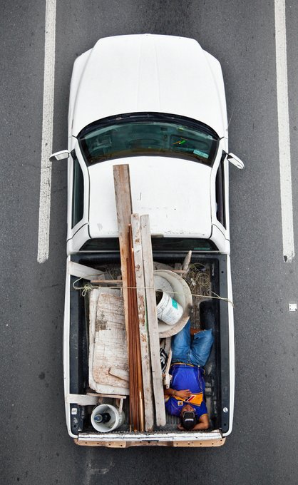 An aerial overhead view of a white pickup truck parked in a lot, loaded with construction materials including wood planks, buckets, and coiled rope in its bed.