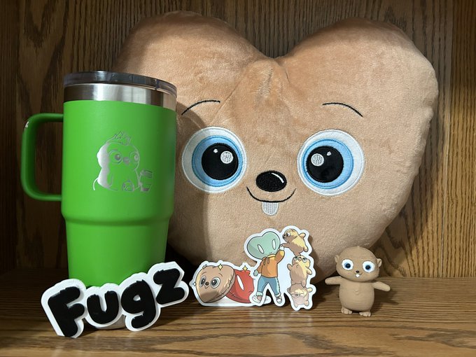A display of 'Fugz' merchandise features a green insulated tumbler, a heart-shaped plush pillow with big eyes, character stickers, and a small brown figurine on a wooden surface.