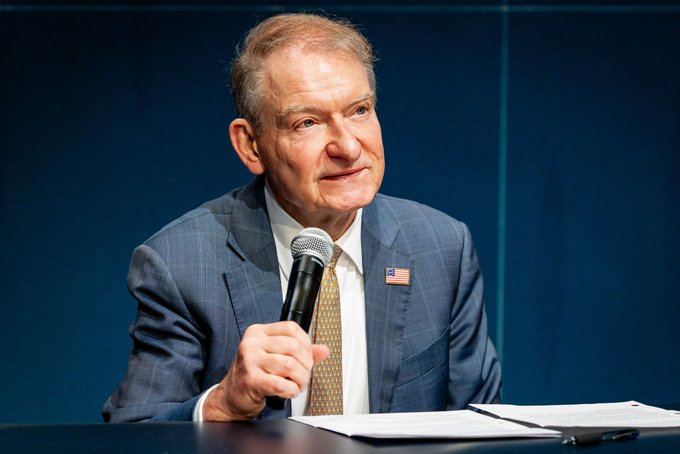 A man in a blue checkered suit holding a microphone, wearing an American flag pin on his lapel, sitting at a table with papers during what appears to be a press conference or panel discussion.