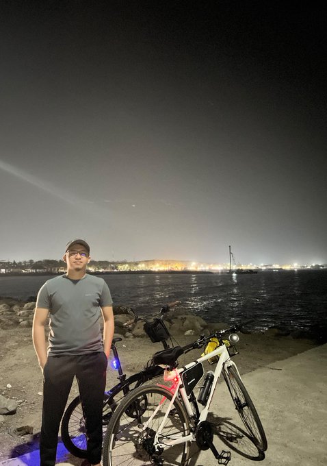 A nighttime photo of a man in a gray t-shirt and cap standing next to two bicycles by a waterfront, with city lights visible across the bay.