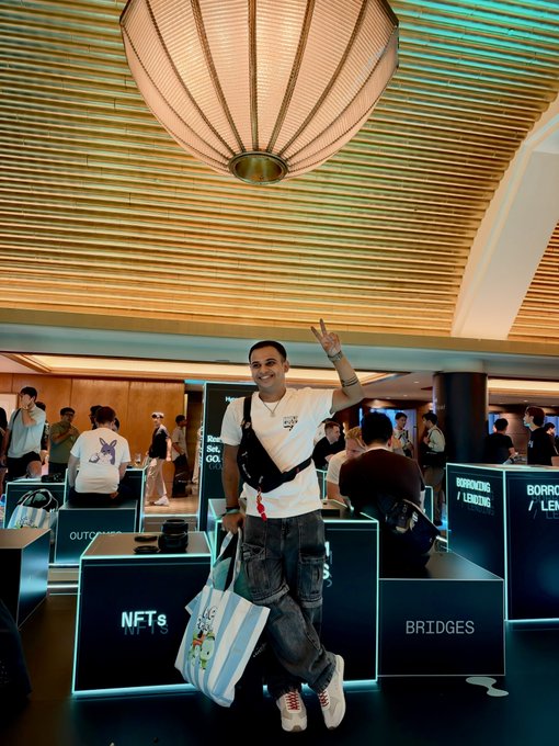 A smiling man in a white shirt poses cheerfully with a peace sign in front of displays labeled "NFTS" and "BRIDGES" at what appears to be an NFT conference. He holds a tote bag while standing amid illuminated booth signage, embodying the crypto/NFT community vibe of the event.