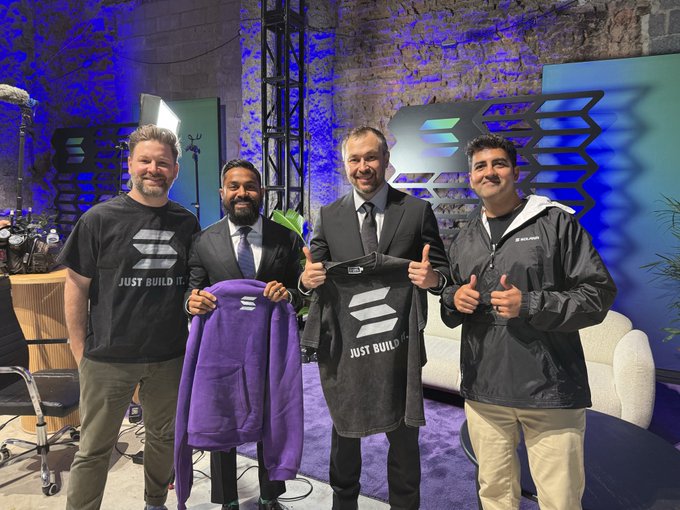 Four men pose for a photo on a stage with purple lighting and geometric Solana branding, holding up apparel displaying the Solana logo and the slogan "JUST BUILD IT." The image ties to the post "Spaces in their faces • One project you should be bullish which I am is @solanaspaces," highlighting a bullish endorsement of the Solana Spaces project through its team members and branded gear.