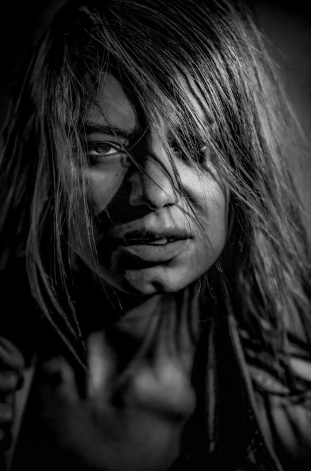 A dramatic black-and-white portrait of a person with long, messy hair partially obscuring their face, looking intensely toward the camera with one hand raised.