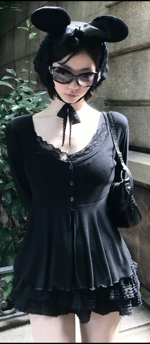 A woman with short black hair wearing a black dress with lace trim, sunglasses, and a black mouse-ear headband, holding a black handbag and standing outdoors near greenery.