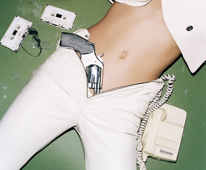 A surreal photograph shows a person's torso with a revolver tucked into the waistband of unzipped white pants, alongside scattered cassette tapes and a telephone on a green surface.