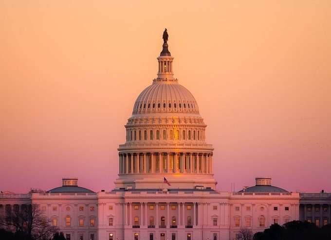 The United States Capitol Building is bathed in a warm sunset glow, its iconic white dome and Statue of Freedom silhouetted against a gradient sky of peach and pink, with an American flag visible on the facade. The image visually anchors the post's focus on the upcoming Senate return and CLARITY Act momentum, symbolizing the legislative developments driving cryptocurrency policy forward in Washington.