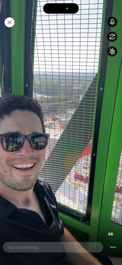 A man wearing sunglasses smiles inside a Ferris wheel cabin, with an aerial city view visible through the grated window. The text "86" appears at the bottom of the screen, and a message field reads "Say something..." — fitting with the post's optimistic "$HBAR going to .05" sentiment about reaching a small price milestone.