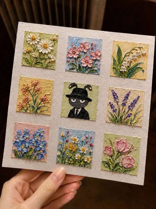 A hand holds a handmade canvas featuring nine small, textured paintings, including floral designs and a central figure of a dog in a suit and hat.