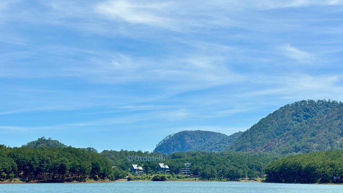 A scenic landscape photograph featuring a lake in the foreground, lush green forested mountains in the background, and a bright blue sky with wispy clouds, watermarked with '@0xdateh'.