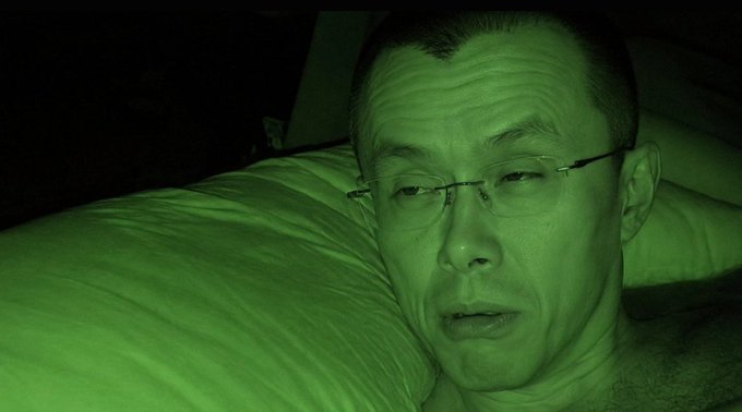 A nighttime selfie of a man wearing glasses, lying on a pillow, with the entire image tinted green, likely due to night vision camera mode.
