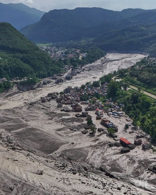 An aerial view of massive flood/landslide devastation in a mountainous village, with a wide debris-covered riverbed and destroyed homes.