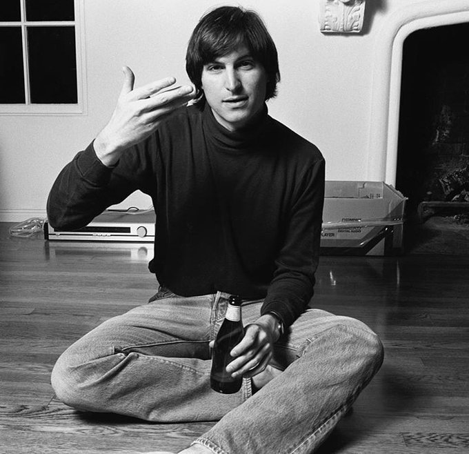 A black-and-white photo of a young Steve Jobs sitting cross-legged on a hardwood floor, holding a bottle of beer and gesturing with his other hand, with Apple-era boxes stacked in the background. The image pairs with the post as a nostalgic nod to Jobs' legacy of making technology more accessible—mirroring the message about increasing ease of engagement with AI and web3.