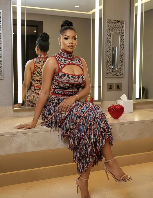 A woman poses confidently in a vibrant, tribal-patterned two-piece outfit featuring fringe details, seated on a marble counter in a modern bathroom with a heart-shaped decoration beside her, exuding a stylish and uplifting energy perfect for a cheerful "Good Morning CT, Happy Thursday" greeting.