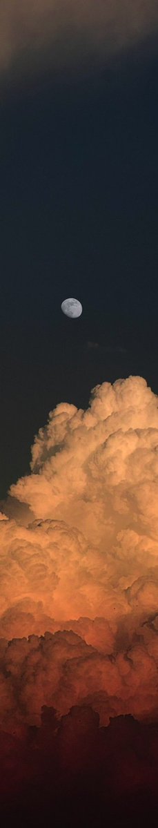 A vertical photograph showing a gibbous moon in a dark night sky above large, billowing clouds illuminated with orange and pink tones.