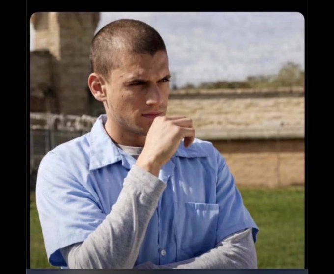 A young man with a buzz cut wearing a light blue button-up shirt poses thoughtfully with his hand near his chin outdoors.