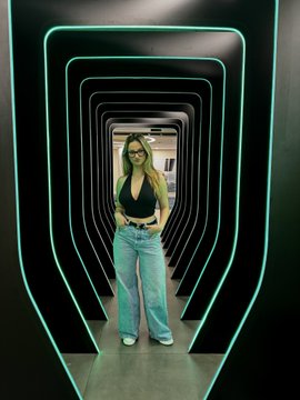 A woman in a black top and light blue jeans stands inside a futuristic tunnel lined with glowing green neon frames.