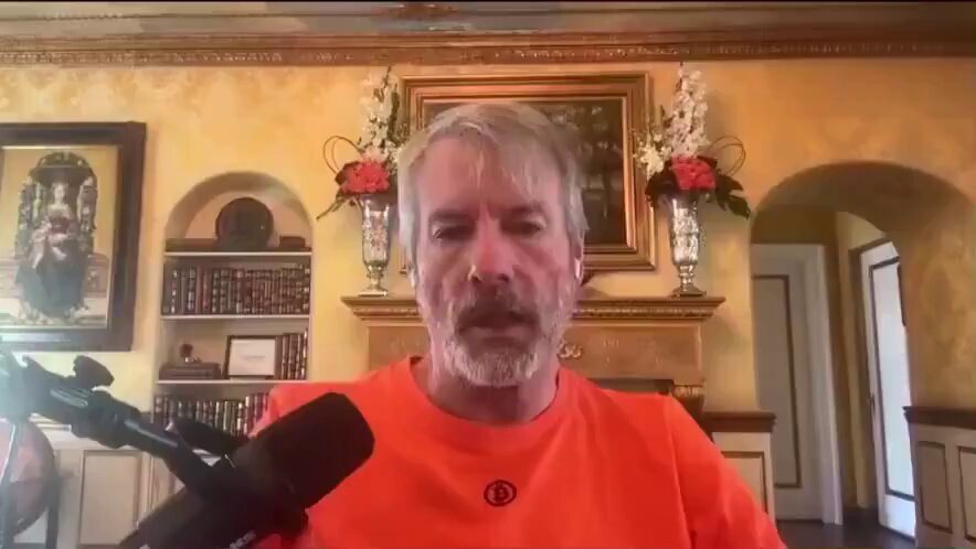 The image features a man wearing an orange shirt standing in front of a fireplace. The photo appears to be part of a podcast or online video format where he is addressing listeners. In his presentation, Michael Saylors talks about Bitcoin's value and its potential for growth. He confidently declares that the cryptocurrency can reach a price of $1 and encourages people to invest in it as a means of gaining substantial wealth. The image captures the man's expression and posture, while the background includes elements like potted plants and vases, adding ambiance to the setting.
