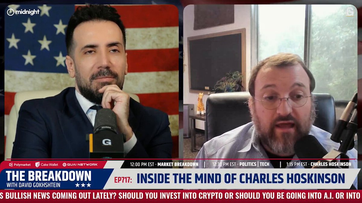 The image shows two men sitting at a table with microphones, appearing to be having a conversation or participating in an interview. The background features an American flag, which suggests that this could be taking place in the United States. The focus of the image is on the two men and their discussion, rather than any specific details about the content displayed on the screen behind them.