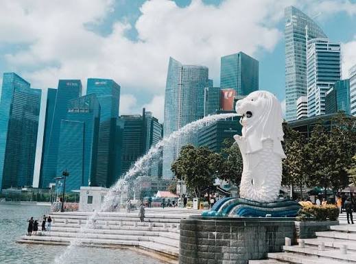 The iconic white Merlion statue of Singapore spouts water from its mouth against a backdrop of towering skyscrapers in the city's financial district.
