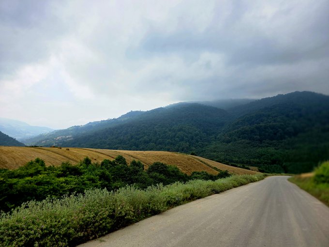 A scenic landscape view of a winding road cutting through mountains under an overcast sky, with forested hills and open fields.