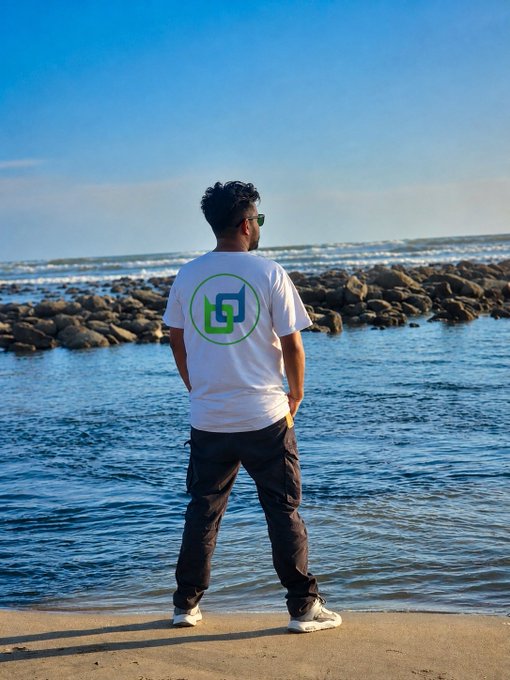 A man viewed from behind stands on a rocky shore looking at the ocean, wearing a white t-shirt with a green circular logo.