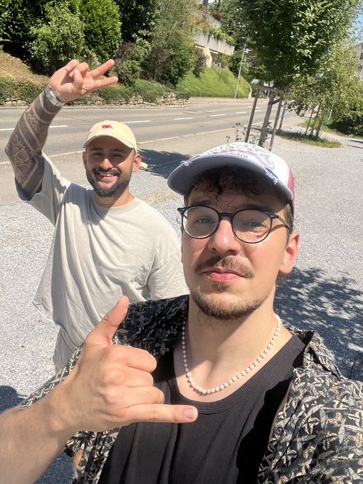 Two men pose for a selfie outdoors on a sunny day; one in front wearing glasses and a patterned cap gives a shaka sign, while the other behind in a beige cap raises a peace sign.