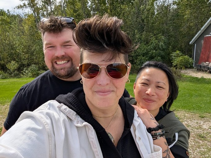 A selfie of three people outdoors on a sunny day, with a person in the center wearing aviator sunglasses and a white jacket, flanked by a smiling man with sunglasses on his head and a woman with dark hair.