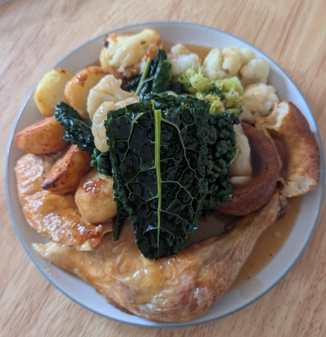 A hearty roast chicken dinner is served in a bowl, featuring golden roasted potatoes, steamed cauliflower, broccoli, savoy cabbage leaves, and Yorkshire puddings drenched in rich gravy. The image perfectly embodies the "Winner Winner Chicken Dinner" saying, showcasing a classic British Sunday roast that looks well worth celebrating.